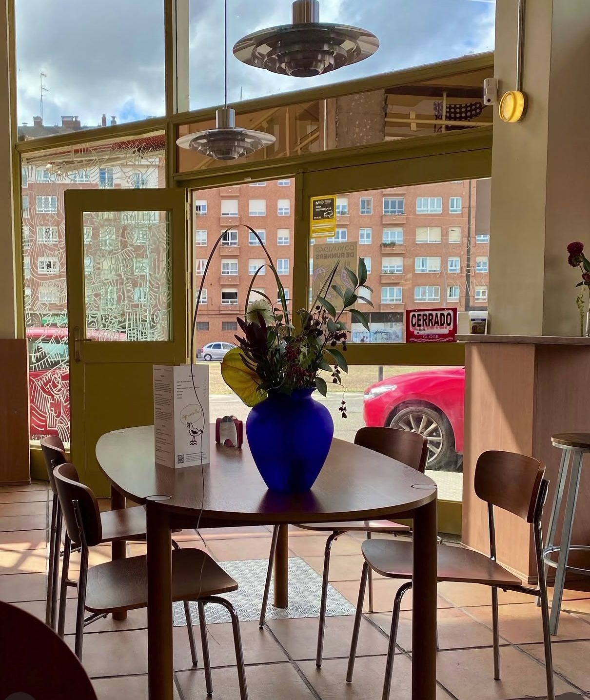 Sala del café con lámparas colgantes, mesas de madera y el jarrón azul bajo los ventanales